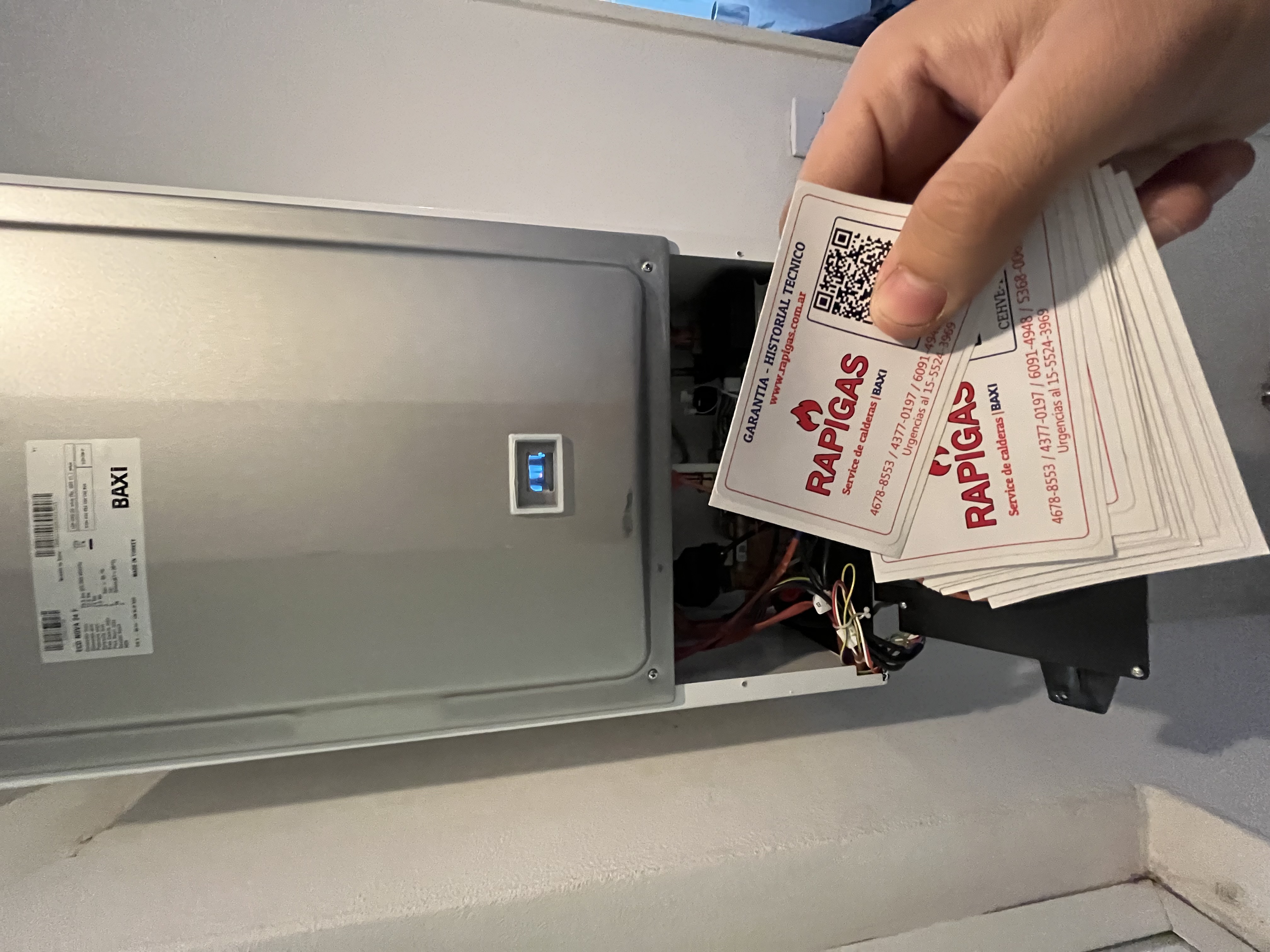 Mano sosteniendo tarjetas de servicio RAPIGAS frente a una caldera Baxi